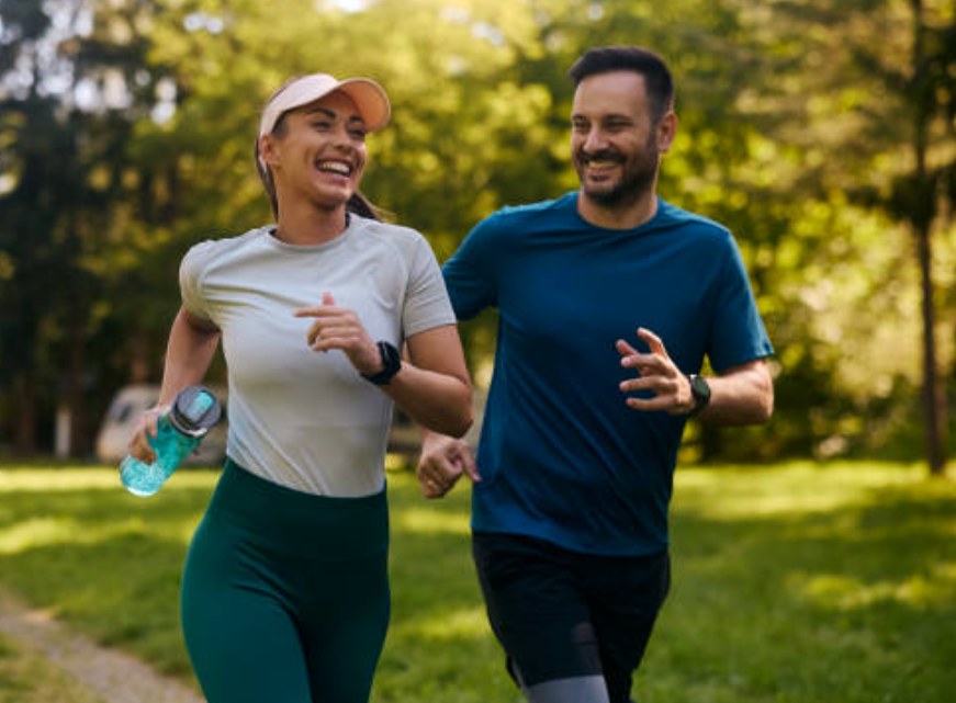 A smiling couple jogging together through a sunlit park, both wearing fitness trackers